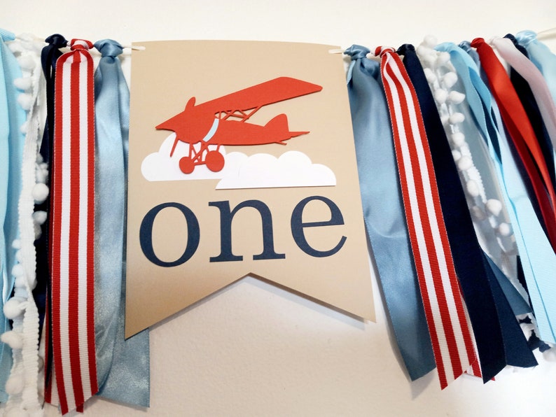 K&ouml;nnte beinhalten: Ein braunes Banner mit einem roten Flugzeug und wei&szlig;en Wolken, das Wort "eins" in blauen Buchstaben und eine Girlande aus blauen, wei&szlig;en und roten B&auml;ndern.