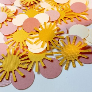 May include: A collection of paper confetti in various shapes and colors. The confetti includes gold sun shapes, along with pink and cream-colored circles. The paper has a textured appearance, and the confetti is scattered across a white surface.