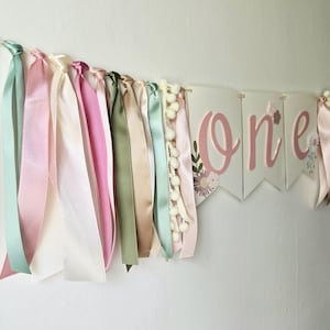 May include: A pastel-colored banner with the word "one" in pink script, decorated with floral accents. The banner is adorned with ribbons in shades of pink, green, and cream, and small white pom-poms. Perfect for a first birthday celebration.