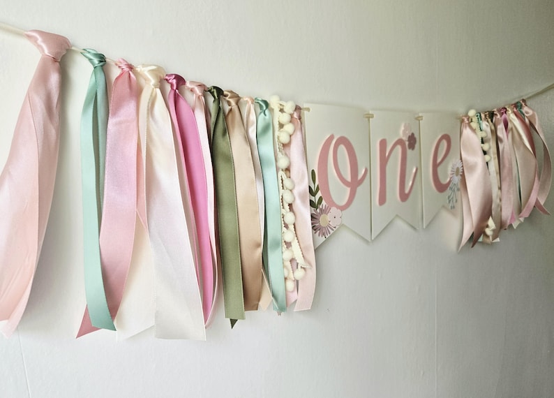 May include: A pastel-colored banner with the word "one" in pink script, decorated with floral accents. The banner is adorned with ribbons in shades of pink, green, and cream, and small white pom-poms. Perfect for a first birthday celebration.
