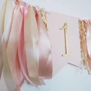 May include: A pink banner with a gold glitter number 1, adorned with ribbons in shades of pink and cream, and small pom-poms. This festive decoration is perfect for a first birthday celebration.