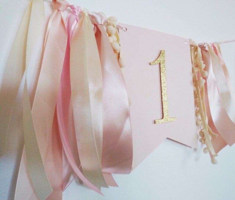 May include: A pink banner with a gold glitter number 1, adorned with ribbons in shades of pink and cream, and small pom-poms. This festive decoration is perfect for a first birthday celebration.
