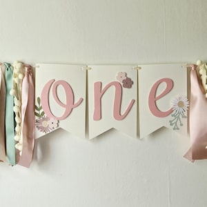 May include: A banner with the word "one" in pink script, flanked by colorful ribbons and pom-poms. The ribbons are in shades of pink, cream, and green. Floral accents are on the banner. The banner is strung on a white string.