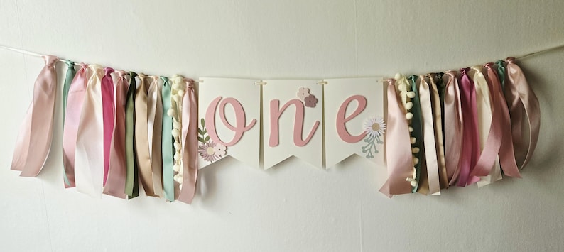 May include: A banner with the word "one" in pink script, flanked by colorful ribbons and pom-poms. The ribbons are in shades of pink, cream, and green. Floral accents are on the banner. The banner is strung on a white string.
