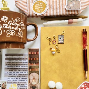 May include: A brown ceramic mug with white mushroom illustrations and the text "Give Thanks" sits beside a yellow notebook, a red pen, and several small metal charms including a key, a flower, a bookshelf, a mushroom, and a leaf.