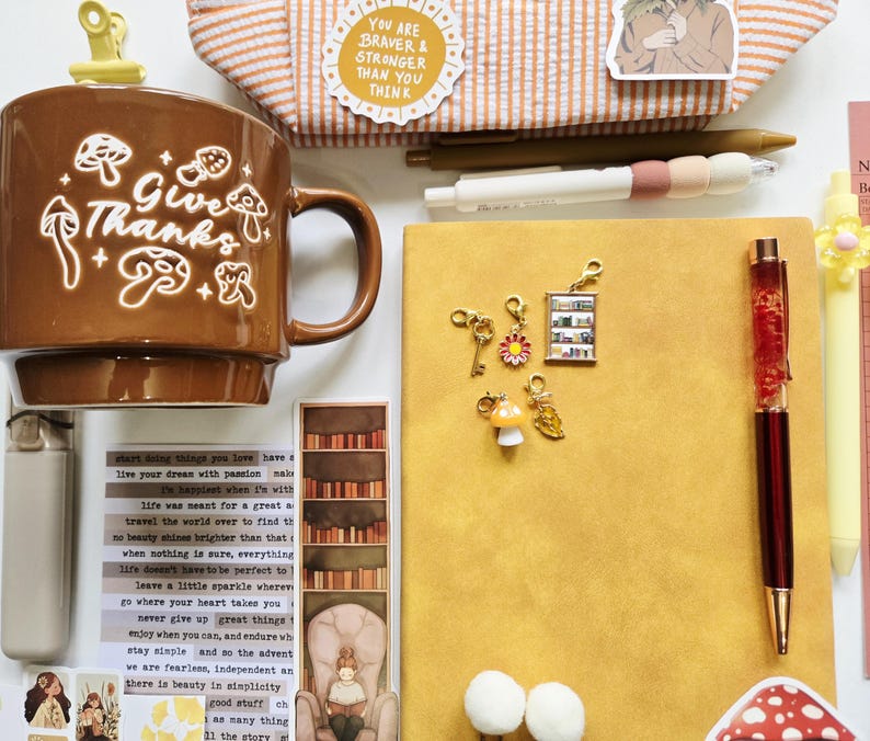 May include: A brown ceramic mug with white mushroom illustrations and the text "Give Thanks" sits beside a yellow notebook, a red pen, and several small metal charms including a key, a flower, a bookshelf, a mushroom, and a leaf.