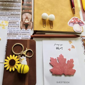 May include: A collection of stationery items including a yellow notebook, a brown notebook, a sunflower and bee keychain, a red maple leaf sticky note labeled feng ye, a mushroom sticker, white pom-pom paper clips, and various decorative paper bookmarks.
