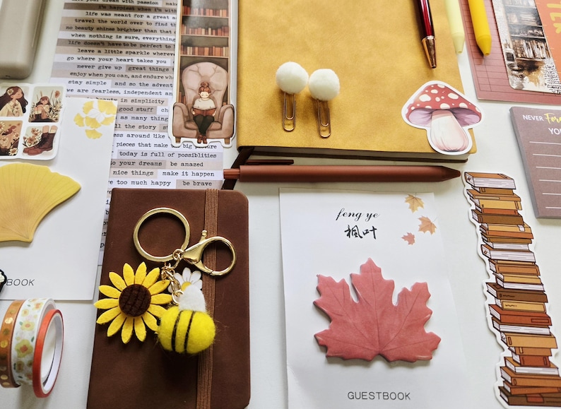 May include: A collection of stationery items including a yellow notebook, a brown notebook, a sunflower and bee keychain, a red maple leaf sticky note labeled feng ye, a mushroom sticker, white pom-pom paper clips, and various decorative paper bookmarks.