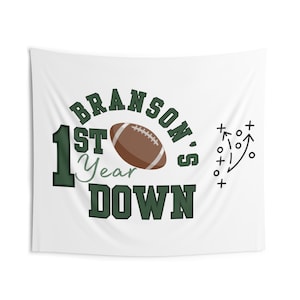 May include: White wall tapestry with the text "BRANSON'S 1ST YEAR DOWN" in green and brown. A brown football graphic is in the center. A black and white football play diagram is on the right side.