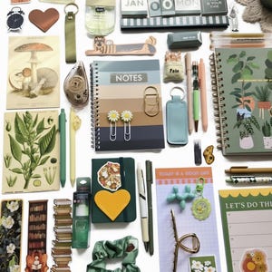 May include: A collection of stationery items including a spiral-bound notebook with a plant print, a gradient-colored notes journal, a daily planner, various pens, daisy-shaped paper clips, a green scrunchie, stickers, and decorative bookmarks.