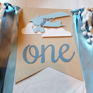 Puede incluir: Una pancarta de papel azul y marr&oacute;n con la palabra "one" en letra azul. La pancarta tiene un avi&oacute;n de papel azul con nubes blancas y cintas azules y doradas.