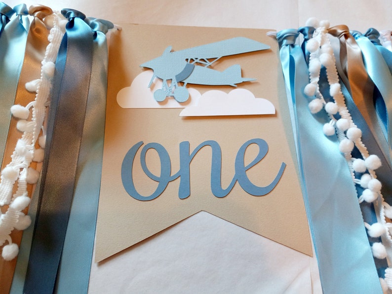 Puede incluir: Una pancarta de papel azul y marr&oacute;n con la palabra "one" en letra azul. La pancarta tiene un avi&oacute;n de papel azul con nubes blancas y cintas azules y doradas.