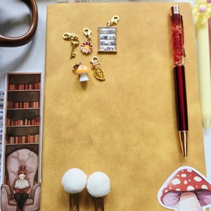May include: A mustard yellow notebook featuring five small gold-toned charms including a key, a flower, a bookshelf, a mushroom, and a leaf. The notebook includes a red pen, two white pom-pom paper clips, and a mushroom sticker on the cover.