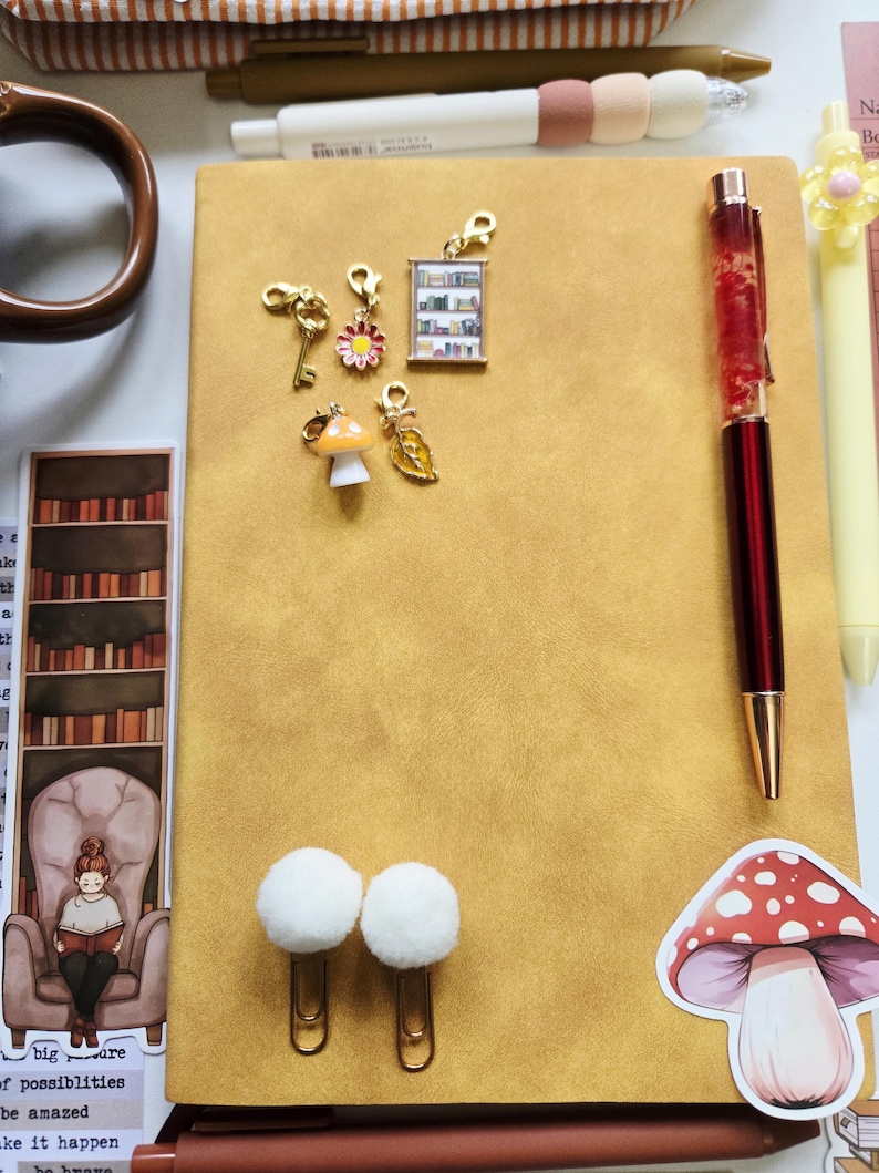 May include: A mustard yellow notebook featuring five small gold-toned charms including a key, a flower, a bookshelf, a mushroom, and a leaf. The notebook includes a red pen, two white pom-pom paper clips, and a mushroom sticker on the cover.