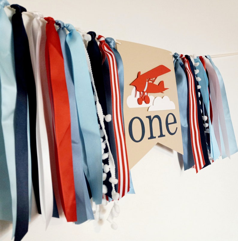 K&ouml;nnte beinhalten: Ein braunes Banner mit einem roten und wei&szlig;en Flugzeug, Wolken und dem Wort "one". Das Banner ist mit blauen, roten und wei&szlig;en B&auml;ndern verziert.