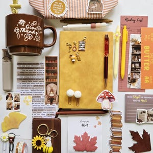 May include: A collection of stationery items including a brown mug with mushroom graphics, a yellow notebook, pens, stickers, bookmarks, paper clips, and a small brown journal. The items feature various illustrations of books, leaves, and flowers.
