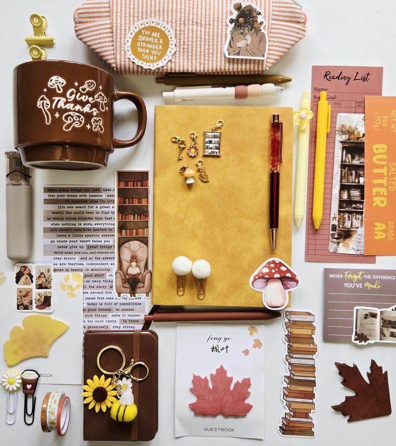 May include: A collection of stationery items including a brown mug with mushroom graphics, a yellow notebook, pens, stickers, bookmarks, paper clips, and a small brown journal. The items feature various illustrations of books, leaves, and flowers.