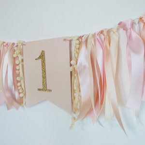 May include: A pink banner with a gold glitter number 1, adorned with ribbons and pom-poms. The ribbons are in shades of pink and cream, creating a festive and celebratory decoration, perfect for a first birthday.