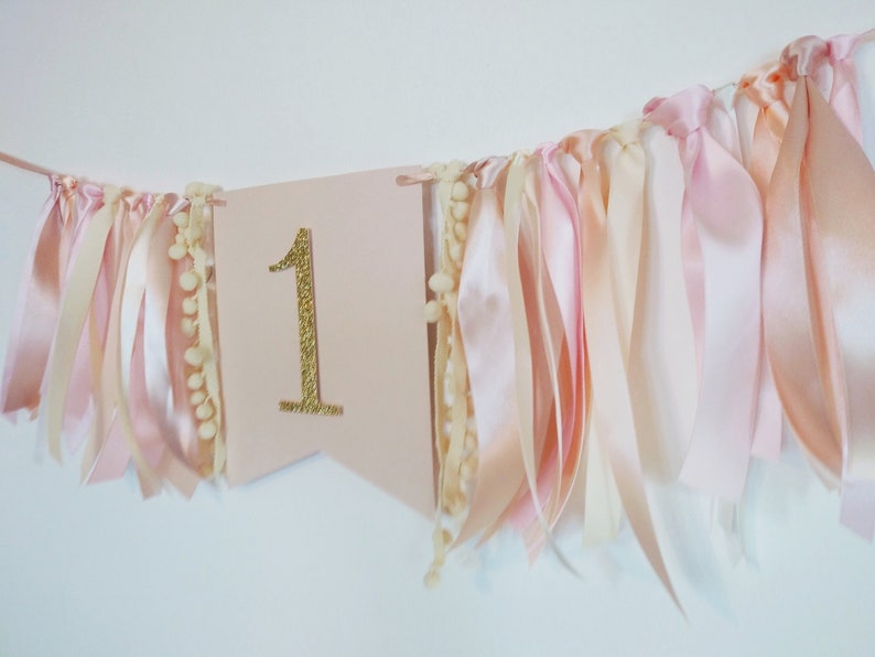 May include: A pink banner with a gold glitter number 1, adorned with ribbons and pom-poms. The ribbons are in shades of pink and cream, creating a festive and celebratory decoration, perfect for a first birthday.