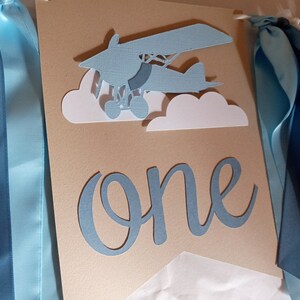 Puede incluir: Una pancarta de papel azul y blanco con la palabra "one" en escritura azul. La pancarta tambi&eacute;n presenta un avi&oacute;n de papel azul con nubes blancas. La pancarta est&aacute; decorada con cintas azules.