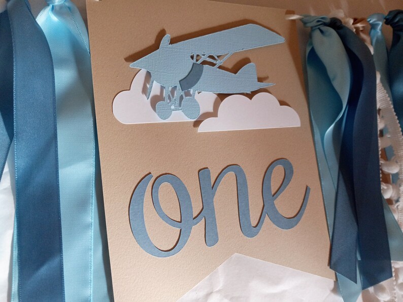 Puede incluir: Una pancarta de papel azul y blanco con la palabra "one" en escritura azul. La pancarta tambi&eacute;n presenta un avi&oacute;n de papel azul con nubes blancas. La pancarta est&aacute; decorada con cintas azules.