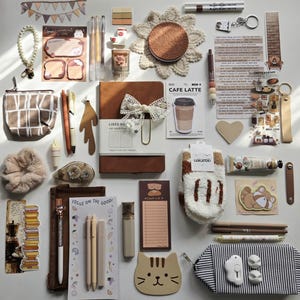 May include: A collection of stationery and lifestyle items including a brown notebook, pens, a pencil case, a cat-shaped notepad, fuzzy socks, a small pouch, stickers, a crochet coaster, and various decorative paper goods arranged on a flat surface.