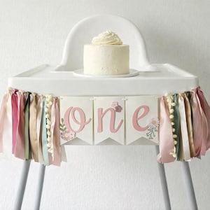 May include: A white high chair with a small cake on top. A banner with the word "one" in pink letters is attached to the front. The banner has ribbons in various colors and small white pom-poms.