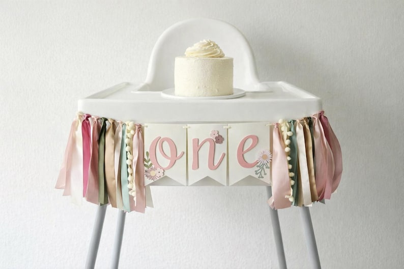 May include: A white high chair with a small cake on top. A banner with the word "one" in pink letters is attached to the front. The banner has ribbons in various colors and small white pom-poms.