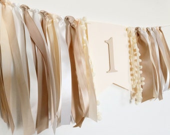 May include: A beige and white fabric banner with the number "1" in the center. The banner is decorated with long, flowing ribbons in shades of beige and white.