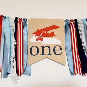 K&ouml;nnte beinhalten: Ein braunes Banner mit dem Wort "one" und einem roten Flugzeug, das in den Wolken fliegt. Das Banner ist mit blauen, roten und wei&szlig;en B&auml;ndern verziert.