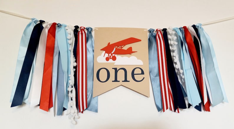 K&ouml;nnte beinhalten: Ein braunes Banner mit dem Wort "one" und einem roten Flugzeug, das in den Wolken fliegt. Das Banner ist mit blauen, roten und wei&szlig;en B&auml;ndern verziert.