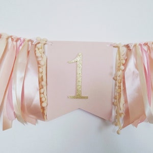 May include: A pink banner with a gold glitter number 1 in the center. The banner is decorated with pink and cream-colored ribbons and small cream-colored pom-poms. The banner is strung on a thin pink string.