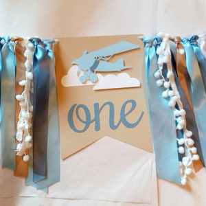 Puede incluir: Una pancarta de papel marr&oacute;n con la palabra "one" en escritura azul. Un avi&oacute;n de papel azul con nubes blancas est&aacute; por encima de la palabra. La pancarta est&aacute; decorada con cintas azules, tostadas y blancas.