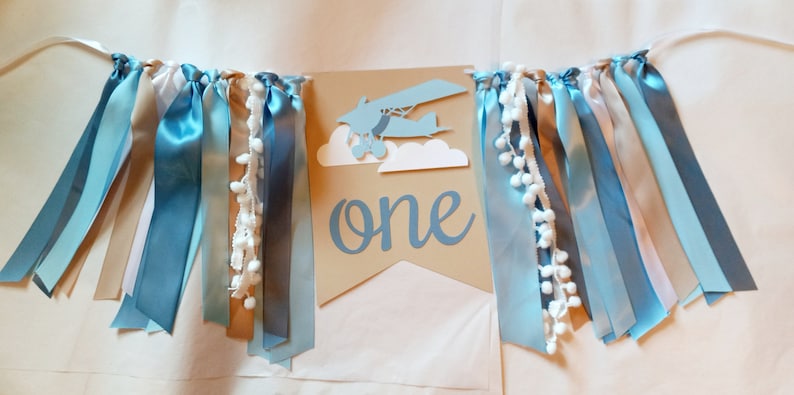 Puede incluir: Una pancarta de papel marr&oacute;n con la palabra "one" en escritura azul. Un avi&oacute;n de papel azul con nubes blancas est&aacute; por encima de la palabra. La pancarta est&aacute; decorada con cintas azules, tostadas y blancas.