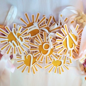 May include: A set of yellow sun-shaped paper cutouts with white rays and white numbers on them. The cutouts are strung together with white and pink ribbon.