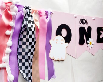 May include: Pink and purple ribbon banner with black felt letters spelling "ONE" and a ghost and spider cutout. The banner is decorated with white pom-poms.