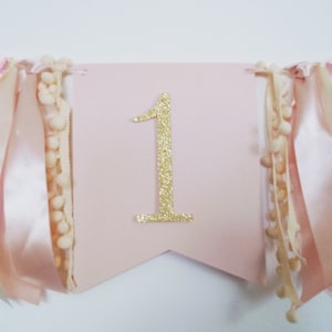 May include: A pink banner with a gold glitter number 1 in the center. The banner is decorated with ribbons in shades of pink and cream, and cream-colored pom-pom trim. Perfect for a first birthday celebration.