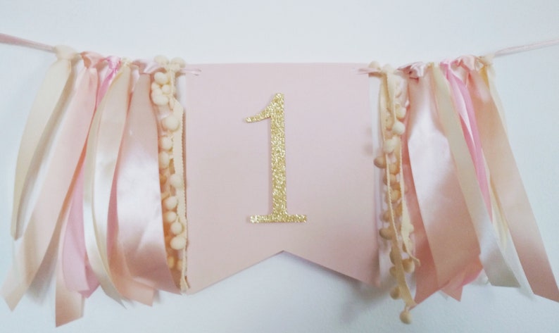 May include: A pink banner with a gold glitter number 1 in the center. The banner is decorated with ribbons in shades of pink and cream, and cream-colored pom-pom trim. Perfect for a first birthday celebration.