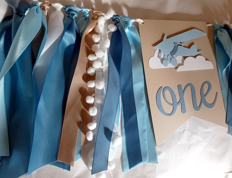 Puede incluir: Una pancarta azul y marr&oacute;n con pompones blancos y cintas. La pancarta tiene un avi&oacute;n azul recortado con nubes blancas y la palabra "one" en azul.