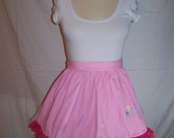 Puede incluir: Una falda rosa con un dobladillo con volantes y un pequeño diseño bordado de globos de colores. La falda se lleva sobre un maniquí blanco.