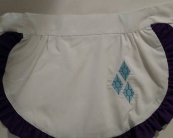Puede incluir: Delantal blanco con ribete de volantes morado y un diseño de diamante azul bordado en la parte delantera.