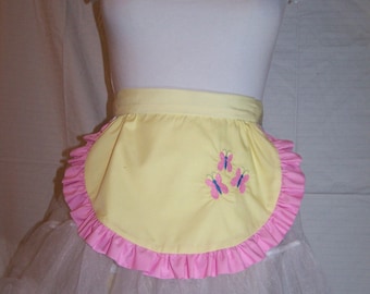 Puede incluir: Un delantal amarillo con un ribete rosa con volantes y tres mariposas bordadas. El delantal se combina con una parte superior blanca y una falda de tul blanca, creando un atuendo caprichoso. El atuendo se muestra en un maniquí.
