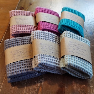 K&ouml;nnte beinhalten: Sechs Waffelpiqu&eacute;-Geschirrt&uuml;cher in verschiedenen Farben, darunter rosa, blau, grau und beige. Jedes Tuch hat ein braunes Papier-Etikett mit handschriftlichem Text in deutscher Sprache, der das Material, die Gr&ouml;&szlig;e und die Waschhinweise angibt.