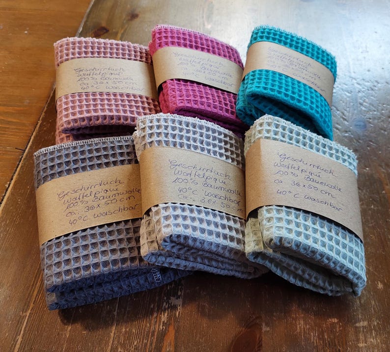 K&ouml;nnte beinhalten: Sechs Waffelpiqu&eacute;-Geschirrt&uuml;cher in verschiedenen Farben, darunter rosa, blau, grau und beige. Jedes Tuch hat ein braunes Papier-Etikett mit handschriftlichem Text in deutscher Sprache, der das Material, die Gr&ouml;&szlig;e und die Waschhinweise angibt.