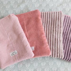 Può includere: Quattro pochette in velluto a coste nei toni del rosa e del viola sono disposte su una superficie a fantasia. Le pochette hanno una forma rettangolare con texture a coste verticali. Una pochette ha un'etichetta con la scritta "made by Pearly Sue."