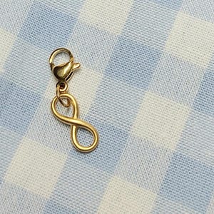 Pode incluir: Um berloque dourado com um símbolo de infinito. O berloque está preso a um fecho lagosta. O berloque está sobre um fundo xadrez azul e branco.