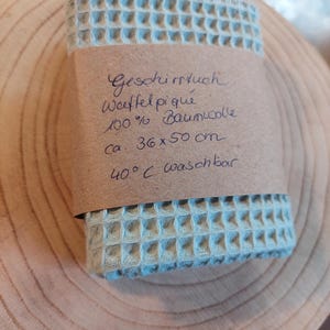K&ouml;nnte beinhalten: Ein hellblaues Waffeltuch aus 100% Baumwolle. Das Tuch ist gefaltet und in braunes Papier eingewickelt, auf dem der Text "Geschirrtuch Waffel piqu&eacute; 100% Baumwolle ca. 36 x 50 cm 40&deg;C waschbar" steht.