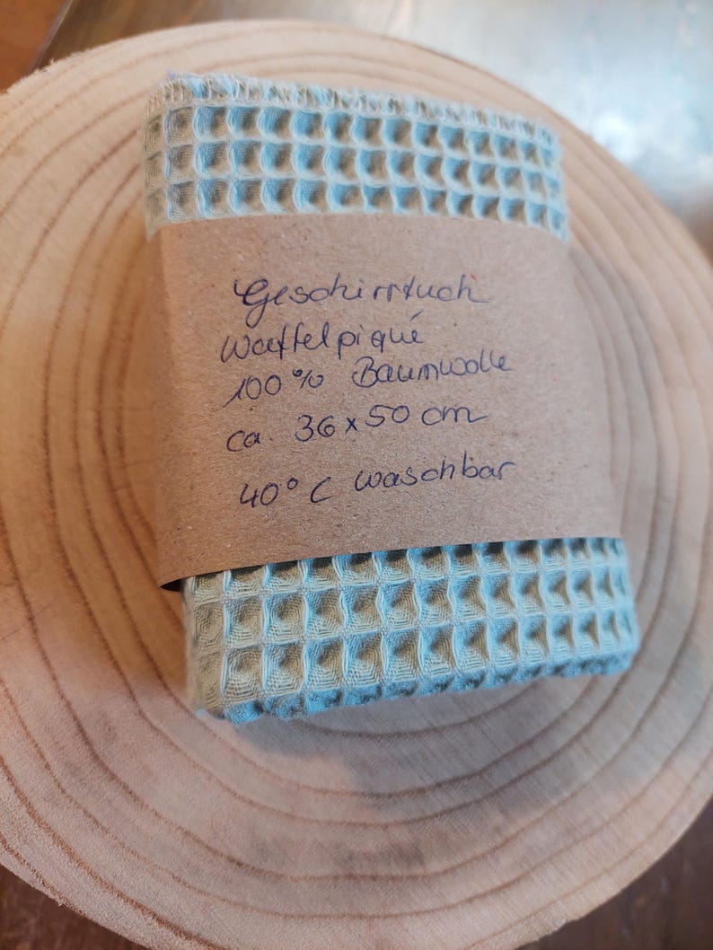 K&ouml;nnte beinhalten: Ein hellblaues Waffeltuch aus 100% Baumwolle. Das Tuch ist gefaltet und in braunes Papier eingewickelt, auf dem der Text "Geschirrtuch Waffel piqu&eacute; 100% Baumwolle ca. 36 x 50 cm 40&deg;C waschbar" steht.