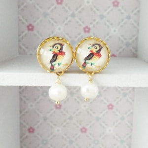 May include: Gold tone earrings with a white background featuring a cartoon bird image and a pearl drop.