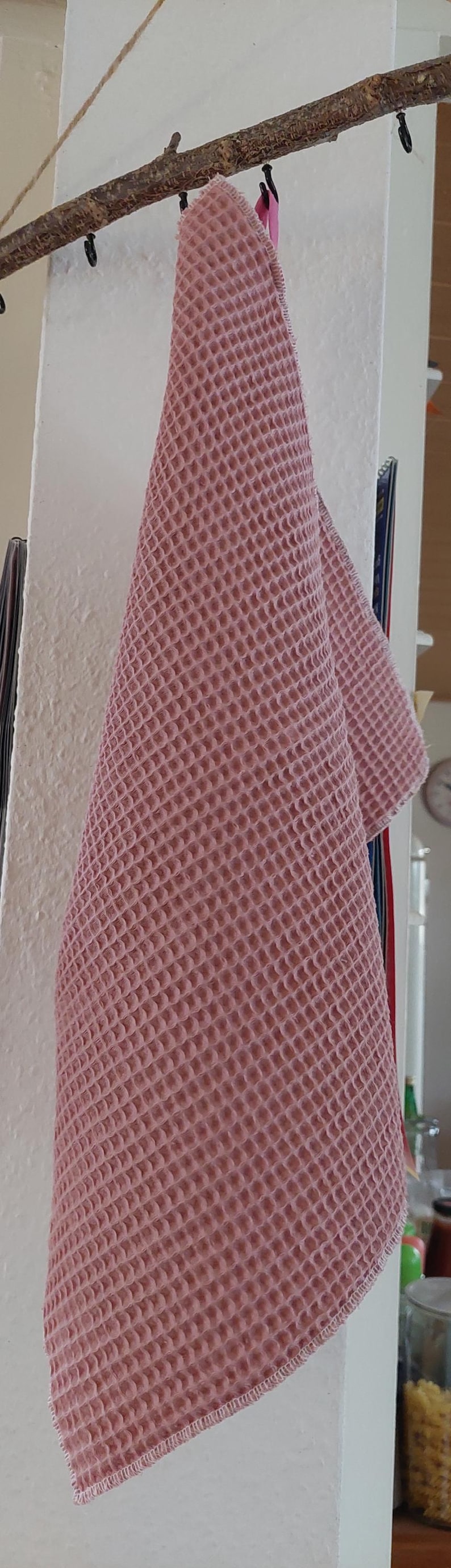 K&ouml;nnte beinhalten: Ein rosa Waffelgewebe-K&uuml;chenhandtuch h&auml;ngt an einem Holzast mit schwarzen Haken.
